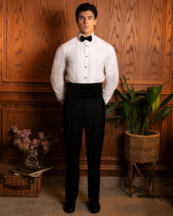 A man wearing a white long-sleeve tuxedo shirt with a black bow tie and black tuxedo pants stands against a wood-paneled wall with decorative plants and a basket.