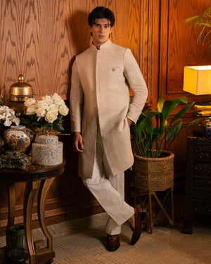 A man wearing a light beige traditional Indian outfit with a long kurta and trousers, posing in a warm, wood-paneled room with decorative plants and a table.