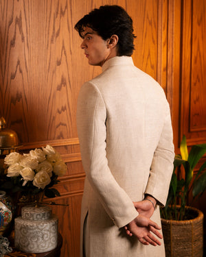 A man wearing a light beige linen tunic jacket with a high collar, shown from the back in a warm, wood-paneled room.