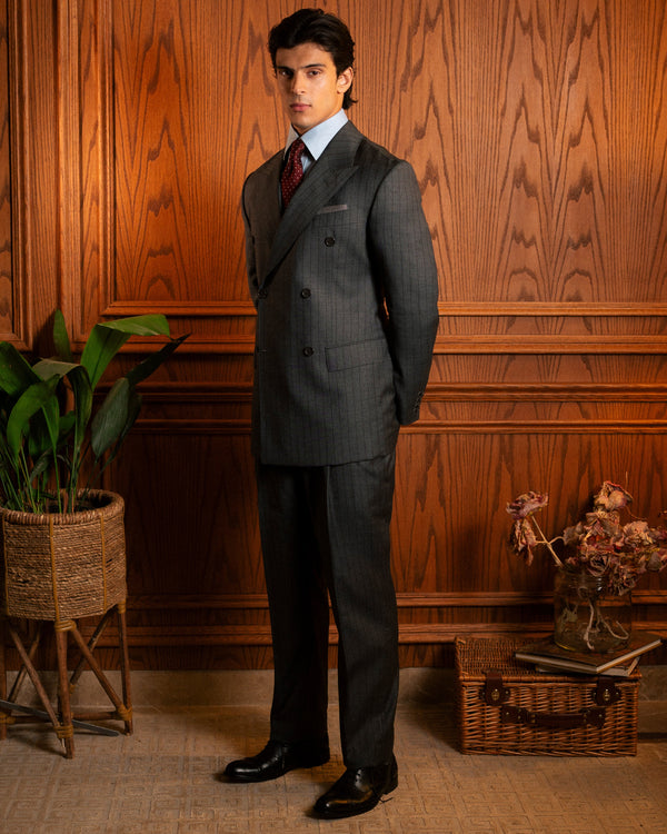 A man wearing a dark gray double-breasted pinstripe suit, white dress shirt, and red tie stands in front of a warm wood-paneled wall. The scene is styled with a potted plant on the left and a small basket with dried flowers on the right.