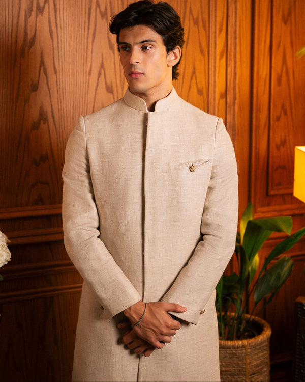A man wearing a light beige linen Nehru jacket with a mandarin collar, standing in front of a warm wood-paneled wall.