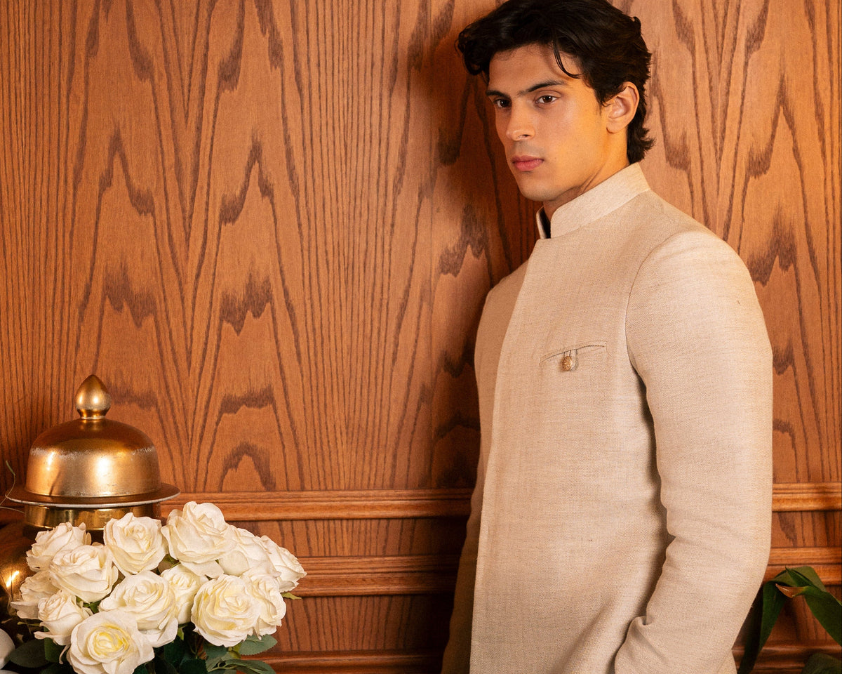 A man wearing a beige long-sleeve kurta stands against a warm wood-paneled wall next to a table with white roses and decorative boxes.