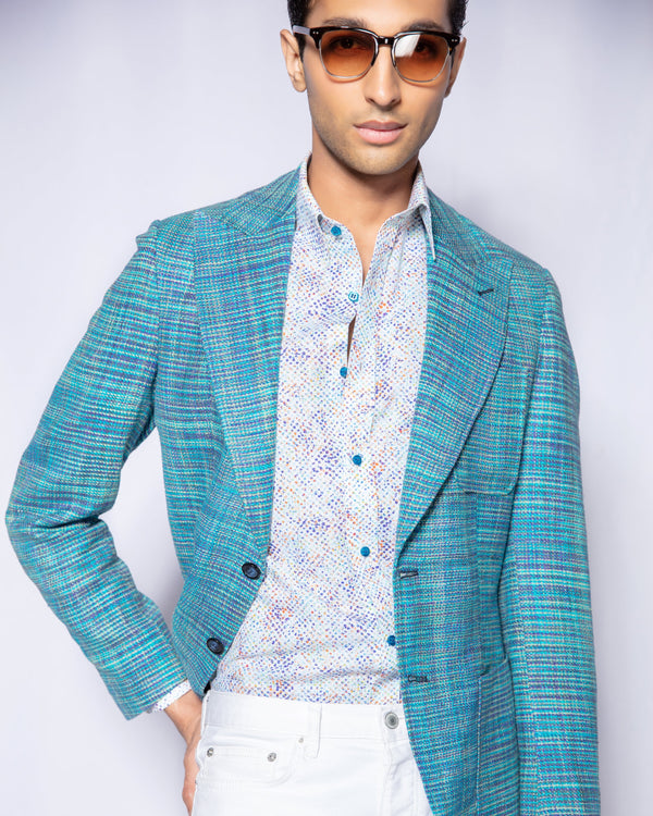 Man wearing a blue checkered blazer over a patterned shirt with white pants on a plain background